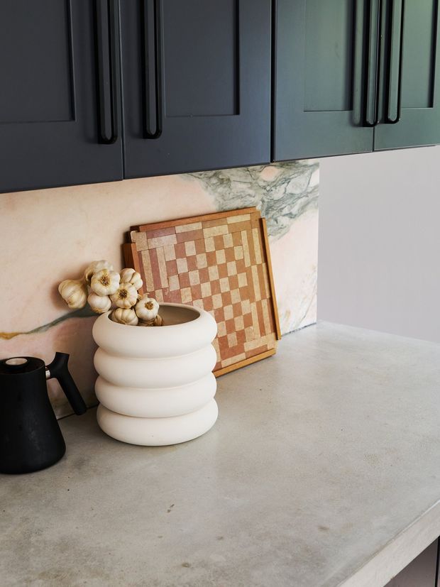 Close-up view of a stylish kitchen benchtop, with dark blue-grey framed cabinetry and elongated black handles above a pale stone or concrete-look work surface. A warm pink-and-cream marble-look splashback forms the backdrop, partially covered by a decorative checkerboard board. A sculptural cream vase and black jug sit on the counter.