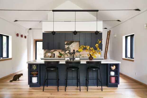 Bright, high-ceilinged kitchen with a large dark island and integrated seating for four. The room features dark blue-gray shaker-style cabinetry, a pale stone benchtop, timber flooring, white walls, a decorative tiled or stone backsplash, black-framed windows, and a linear multi-pendant light over the island. Open shelving at the island ends displays tableware, while a cat and floral arrangement add incidental visual interest.