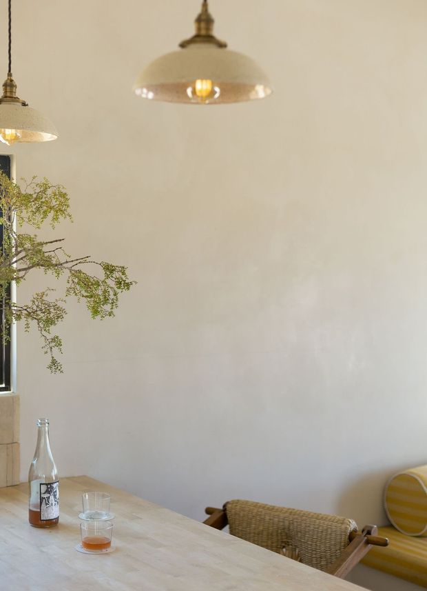 A softly lit dining area with a pale plaster wall, two vintage pendant lights, and a light timber dining table. Woven dining chairs and a striped upholstered chair sit around the table, with a simple glass bottle and drinksware on its surface. A leafy branch and partial window appear at the left edge, creating a relaxed, warm interior atmosphere.