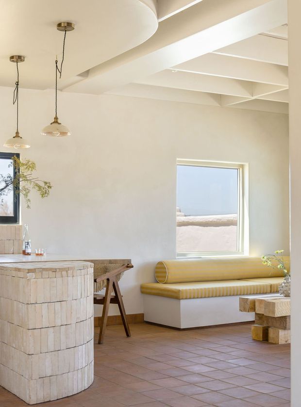 Bright, warm-toned open-plan interior with a curved tiled kitchen island in the foreground, a timber dining table and chairs, and a built-in window seat upholstered in mustard-yellow striped fabric. The space features cream walls, exposed white-painted ceiling beams, pendant lights, terracotta-toned tiled flooring, and a large window with an exterior view. A chunky timber coffee table or side table is partially visible at right.