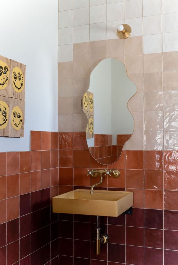 A compact, characterful bathroom corner featuring a wall-mounted mustard-beige basin with exposed brass plumbing and vintage-style brass tapware. The walls are clad in glossy square tiles arranged in bands: pale blush and white tiles above, terracotta tiles through the middle, and deep burgundy tiles below. An organically shaped frameless mirror sits above the basin, reflecting playful yellow artwork, while additional smiley-face artwork is mounted on the adjacent wall. The composition has a bold retro, eclectic feel with warm colours and glossy surfaces.