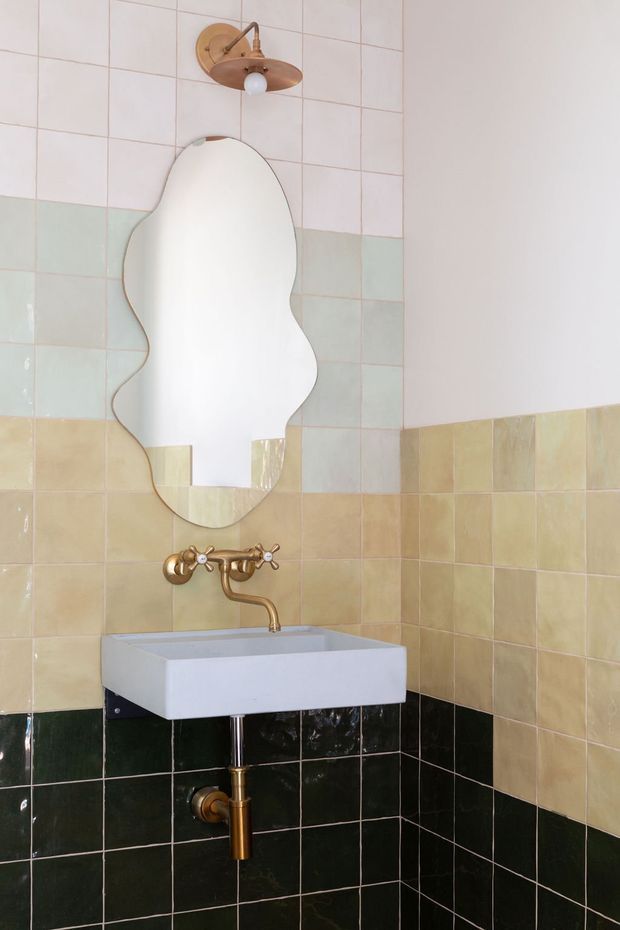 A compact bathroom corner with a wall-mounted rectangular white basin, exposed brass plumbing and vintage-style brass tapware. The walls are tiled in horizontal bands of pale pink, mint green, beige, and dark green square tiles. An irregular organic-shaped mirror and a small copper-toned wall sconce add distinctive decorative details.