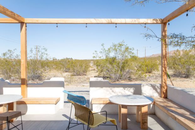 A bright outdoor dining and lounge terrace with built-in white rendered bench seating, timber tables, a woven lounge chair, and a timber pergola frame with festoon lights. The open terrace overlooks a dry desert-like landscape with low scrub and distant hills under a clear blue sky.