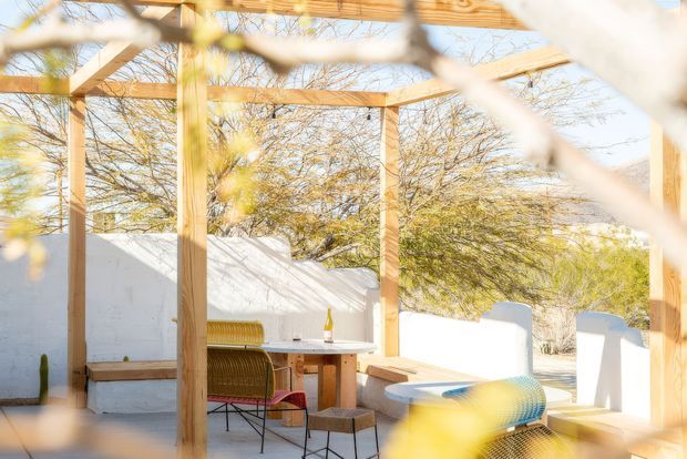 Bright outdoor courtyard dining and lounge area beneath a timber pergola, enclosed by white rendered walls and mature trees. A small dining table with chairs, built-in bench seating, and a curved outdoor lounge seat create a relaxed, sunlit setting.