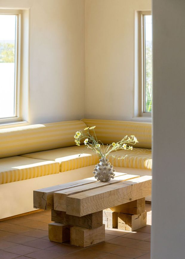 A sunlit interior seating nook with built-in L-shaped banquette seating upholstered in yellow-and-white striped fabric, positioned beneath two tall windows. A chunky reclaimed-looking timber coffee table and a small vase of delicate flowers sit on a terracotta-tiled floor. The space has warm cream walls, abundant natural light, and a simple relaxed aesthetic.
