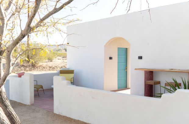 Bright exterior view of a whitewashed, low-rise courtyard or terrace with curved stucco privacy walls, a turquoise-painted door set within an arched opening, and a simple built-in bar counter with a dark red tiled or stone support. Sparse planting and mature trees surround the outdoor sitting area, creating a serene desert-like setting.