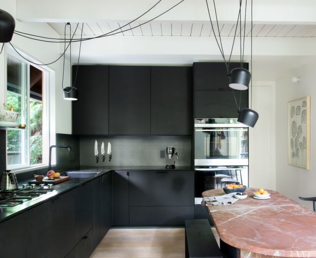 A contemporary black kitchen with matte dark cabinetry, integrated stainless-steel appliances, a dark reflective splashback, and a large veined stone island or dining bench. A timber floor, white ceiling with exposed linear detailing, pendant lights on long black cords, and a window overlooking greenery complete the space. The dining area and framed artwork are visible to the right.