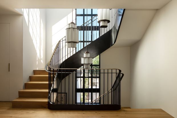 A bright, double-height stair hall with a sculptural curved staircase. Warm timber treads and flooring contrast with dark metal balustrades and a black stair structure. Tall glazed openings bring in abundant daylight, while several suspended geometric pendant lights hang through the stair void. A minimalist white door and walls complete the refined modern interior.