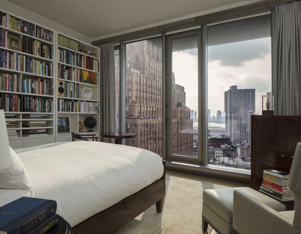 A refined high-rise bedroom with a large upholstered bed, floor-to-ceiling corner glazing, built-in white bookshelves filled with books, and a pair of light armchairs. The room overlooks a dense urban skyline and appears bright but softly muted, with neutral furnishings, dark timber furniture, and full-height grey curtains.