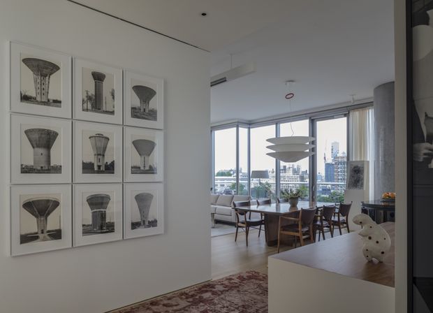 A bright, refined open-plan apartment interior viewed from a hallway or entry area, looking toward a dining space and glazed living area. A gallery wall of framed black-and-white architectural photographs decorates the white wall, while a timber dining table and chairs sit beneath a sculptural pendant light. Floor-to-ceiling corner glazing provides an urban skyline view, and a white built-in surface with decorative objects occupies the foreground.