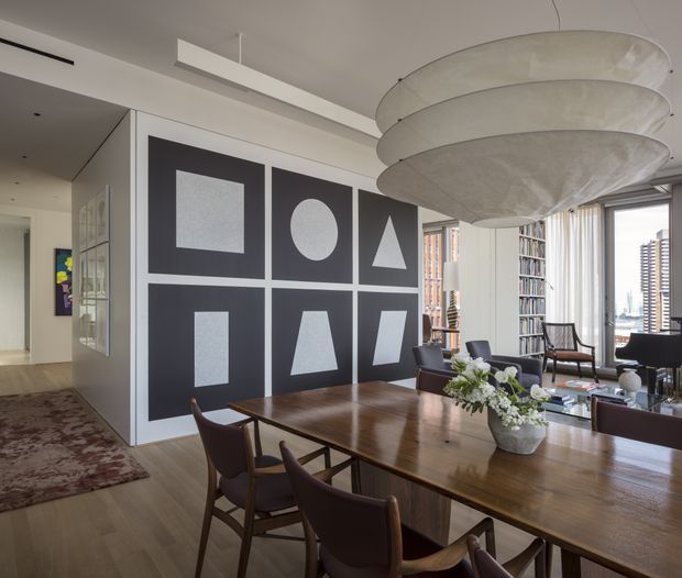 Refined contemporary dining and living area with a large dark geometric feature wall, timber dining table and leather dining chairs. A sculptural layered pendant light hangs above the table, while floor-to-ceiling windows provide city views and abundant natural light. The space includes a small lounge seating area and built-in cabinetry along the rear wall.