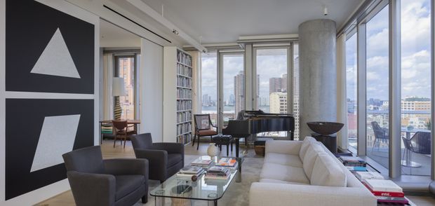 A bright, high-rise apartment living room with floor-to-ceiling glazing and expansive city views. The space features a white sectional sofa, dark lounge chairs, a glass coffee table, a grand piano, extensive built-in shelving, large abstract wall art, and a prominent exposed concrete column. The interior has a refined contemporary urban character with abundant natural light.