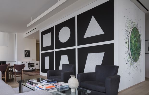 Contemporary open-plan living and dining area with two dark armchairs facing a glass coffee table, a small dining setting and compact kitchen visible in the background. The space features white walls and ceiling detailing, pale timber flooring, and a bold black-and-white geometric feature wall with square, circle and triangle artwork panels. A sculptural circular mural adds visual interest to the adjacent wall.
