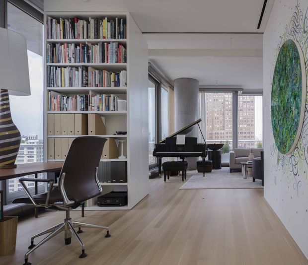A refined high-rise apartment interior with a home office in the foreground and an open living area beyond. The office includes a built-in floor-to-ceiling bookshelf with books and storage binders, a dark ergonomic desk chair, and a desk beside expansive windows. A grand piano sits in the adjoining living space, which has pale timber flooring, neutral furnishings, large windows, and a prominent cylindrical structural column. A large colourful artwork occupies the right wall. The overall space is bright, contemporary, and sophisticated.