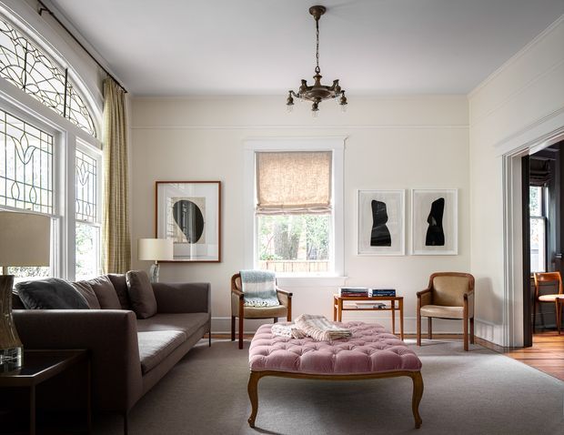 A refined traditional living room with high ceilings, decorative leaded-glass bay windows, ornate chandelier, classic wall mouldings, and a neutral palette. The room contains a dark upholstered sofa, two occasional armchairs, a tufted blush-pink ottoman, framed artwork, and a timber side table. A doorway opens to an adjacent room with additional timber detailing.