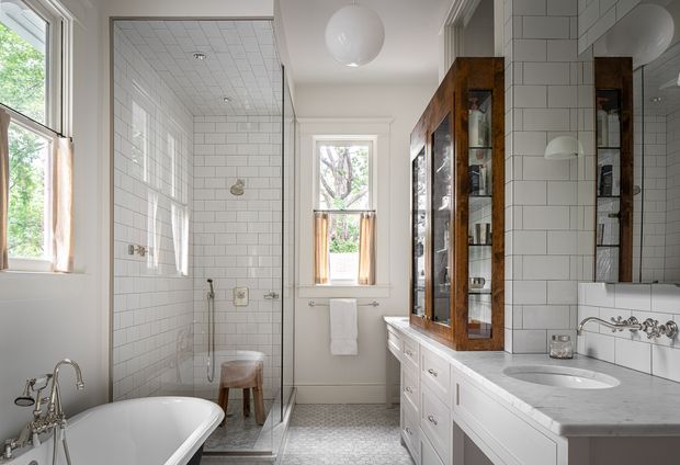 Bright traditional bathroom with a freestanding white bathtub, glass-enclosed tiled shower, single vanity with white cabinetry and a pale stone benchtop, and a tall dark timber mirrored cabinet. White subway wall tiles, patterned floor tiles, timber-trimmed windows, polished chrome fixtures, and a spherical pendant create a refined, airy interior.