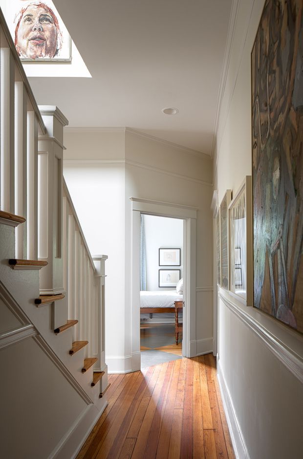Bright, narrow residential hallway beside a staircase, featuring warm timber floorboards, painted decorative wall panelling and trim, a skylight, framed artwork, and a bedroom visible through the doorway at the end. The space has traditional detailing and abundant natural light.