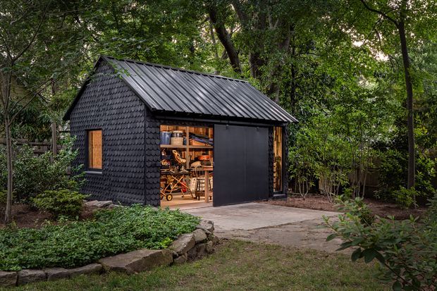 A compact dark-clad standalone shed or studio in a leafy garden setting. The small structure has a pitched corrugated metal roof, textured dark shingle-like wall cladding, warm timber-framed openings, and a large open sliding door revealing workshop storage and equipment inside. A concrete path and low stone-edged garden bed lead to the building.
