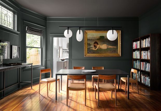 A dark, richly detailed dining room with a long black dining table surrounded by warm timber-and-upholstered chairs. Deep charcoal wall panelling, polished timber flooring, built-in shelving filled with books, framed artwork, pendant globe lights, and tall windows create a moody yet refined interior. A compact sideboard and artwork are visible along the left wall, with a glimpse into an adjoining space through the rear opening.