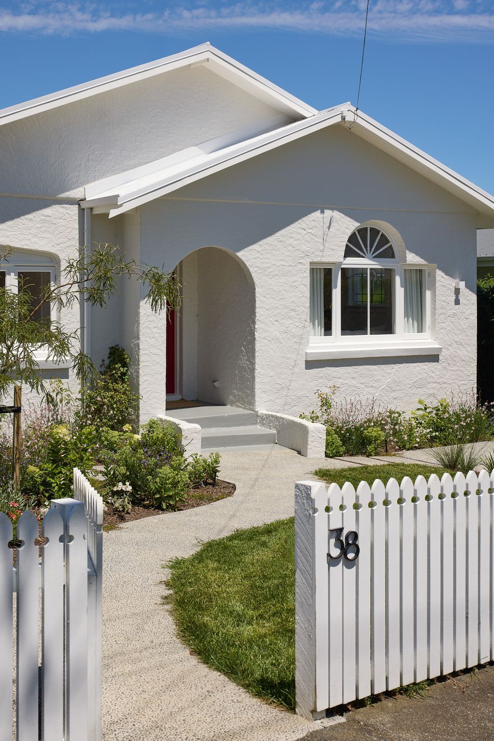 The original white stucco bungalow sits comfortably behind a traditional picket fence and established cottage garden. A gently winding garden path leads visitors to the welcoming front entrance, celebrating the home's heritage character.