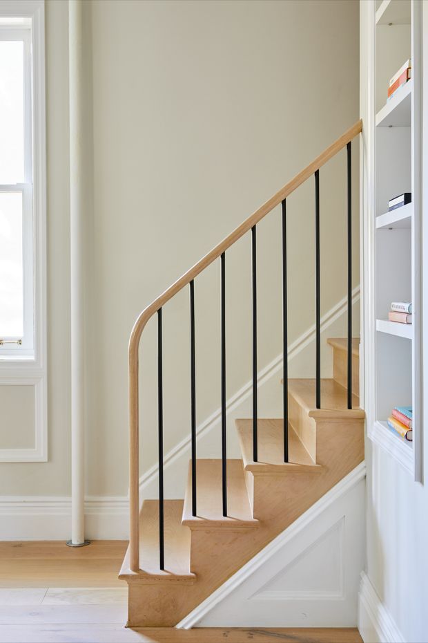 A bright, narrow interior stairway with a crafted timber staircase, curved timber handrail, and slender black metal balusters. The stairs rise beside a white-painted paneled wall and built-in open shelving filled with books. Traditional architectural detailing, including decorative wall panelling, moulded skirting, and white trim, contrasts with the clean contemporary stair balustrade. A tall window brings in natural light.