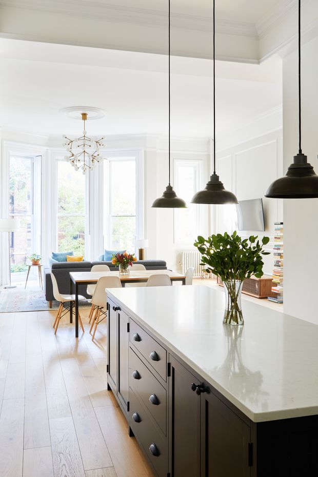 Bright, open-plan kitchen and dining/living space in a period-style home. A large island with dark painted cabinetry and a pale stone-like benchtop dominates the foreground, lit by three black pendant lights. White walls with decorative panel moulding, tall bay-style windows, pale timber flooring, a dining table, and a lounge area create an airy, refined interior.