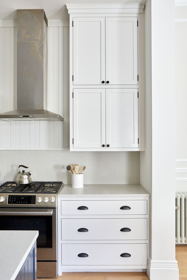 A bright, compact traditional-style kitchen vignette with white Shaker cabinetry, black pulls, a pale stone benchtop, vertical white wall panelling, stainless-steel rangehood and freestanding cooker. A kettle and utensil holder sit on the cooktop and benchtop. Warm timber flooring and classical crown moulding and skirting are visible, with a radiator glimpsed through the opening at right.