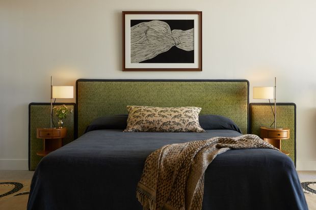 A styled bedroom with a large bed centered against a green upholstered headboard, matching upholstered side panels, and dark navy bedding. A patterned accent cushion and draped textured throw add warmth. Slim bedside tables with table lamps flank the bed, while framed monochrome artwork hangs above. The room has pale neutral walls, carpeted flooring, and a refined, moody yet inviting atmosphere.