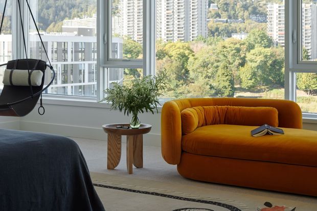 Bright contemporary living space with expansive floor-to-ceiling windows overlooking a leafy urban setting and high-rise buildings. The room features a distinctive mustard-orange curved sofa, a black hanging egg chair, a small timber side table with a potted plant, a dark-covered bed or daybed in the foreground, and a patterned rug. The interior is airy and furnished with rounded, sculptural forms.