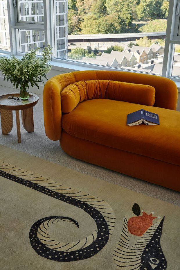 A bright high-rise living room corner with floor-to-ceiling windows overlooking an urban residential setting. The space features a sculptural mustard-orange velvet sofa, a small wooden side table with a glass vase and greenery, and a large beige area rug with bold black and abstract botanical motifs. An open book rests on the sofa, adding a relaxed residential feel.