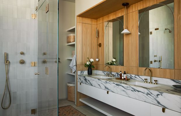 Contemporary bathroom with a glass-enclosed tiled shower on the left and a long double vanity on the right. The vanity features white cabinetry, a dramatic white-and-grey veined stone benchtop and splashback, warm timber wall panelling, large mirrored cabinets, brass-toned fixtures, and open shelving with towels and baskets. The room has a refined, warm modern aesthetic.