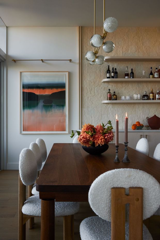 A refined dining room with a dark timber rectangular dining table surrounded by sculptural boucle upholstered chairs. A dramatic multi-globe pendant hangs above the table, while a textured pale stone or ceramic feature wall with open shelves displays bottles and glassware. A framed abstract artwork, floral arrangement, candles, and warm timber flooring complete the sophisticated, warm-toned interior.