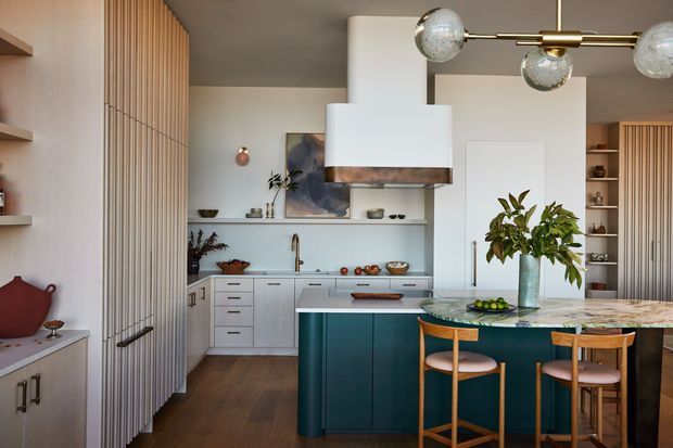 Warm contemporary kitchen with a central teal island, pale cabinetry, a veined stone benchtop, timber bar stools, vertical timber wall panelling, open shelving, and a sculptural brass-and-glass pendant light. The space has a refined mid-century influence with a clear connection between the kitchen and dining area.