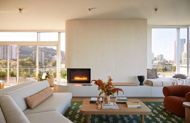 Bright contemporary living room with expansive floor-to-ceiling glazing overlooking a dense urban landscape. The room features a large curved fluted feature wall with an inset fireplace, pale upholstered sofas, a sculptural orange lounge chair, a low timber coffee table, and a patterned green rug. The open, light-filled arrangement has warm neutral finishes and refined modern furnishings.