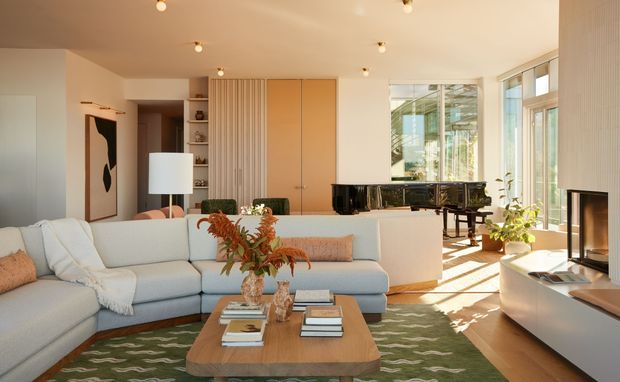 Bright, spacious contemporary living room with a large pale sectional sofa, timber coffee table, sculptural green rug, and a grand piano. Warm timber cabinetry and vertical timber wall detailing frame the rear of the room, while extensive glazing brings in abundant natural light. A fireplace and low built-in media joinery are visible on the right, with indoor plants and layered neutral furnishings creating a refined, inviting atmosphere.