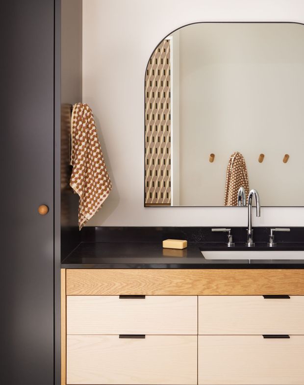 A compact, refined bathroom vanity is shown straight on. The built-in cabinetry has light timber drawer fronts with slim dark pulls, topped by a polished black benchtop and integrated basin. A tall chrome mixer tap sits beneath a large rounded-corner mirror. A patterned hand towel hangs beside a dark vertical cabinet, with a small soap bar on the counter and simple wall hooks reflected in the mirror.