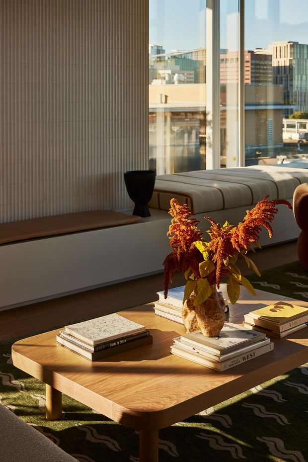 A warmly lit contemporary living room overlooking an urban cityscape through expansive floor-to-ceiling windows. The room features a low upholstered sofa or daybed, a large rounded-corner timber coffee table, sculptural black decorative object, patterned dark rug, and stacks of books with a vase of dried orange foliage. Vertical ribbed wall panelling and a long low built-in ledge add architectural texture.