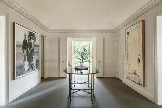 A refined, symmetrical residential entry hall with pale paneled walls, ornate ceiling cornices, dark flooring, and a central glass-topped console table. Large monochrome artwork and a framed map add visual interest, while glazed doors at the far end open toward greenery. The space has a bright, calm, traditional character.