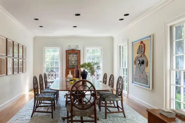 Bright, elegant dining room with a long rectangular table surrounded by ornate dark wood dining chairs, including a prominent wheel-back chair in the foreground. The room has white walls with substantial crown moulding, recessed ceiling lights, tall divided-light windows and French doors, polished timber flooring, a large pale patterned area rug, framed artwork, and a traditional wooden display cabinet. The styling combines classic architectural detailing with eclectic vintage furnishings and art.