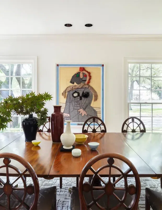 Bright traditional dining room with a large polished dark-wood dining table, matching wheel-back dining chairs, white walls and trim, divided-light windows, recessed ceiling lights, decorative pottery, greenery, and a framed artwork. The room has abundant natural light and a refined, classic character.