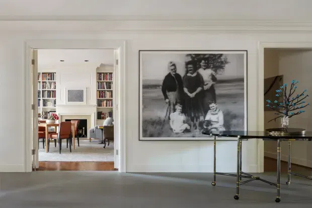 A refined, light-filled interior view of a formal hallway or entry space with pale walls, detailed white architectural trim, and a grey carpeted floor. A large framed monochrome family photograph forms the main wall feature. Through the open doorway, a traditional living room is visible with built-in white bookcases, a fireplace, seating, and a dining table. A glass-topped console table with slender dark metal supports and a decorative branch arrangement occupies the foreground.