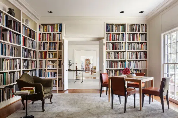 Bright, refined library or reading room with floor-to-ceiling built-in bookshelves on three sides, a small dining or reading table with leather chairs, a classic upholstered armchair, a large neutral rug, and traditional architectural trim. The room opens through a central doorway toward additional interior spaces and receives abundant daylight through a tall multi-pane window.