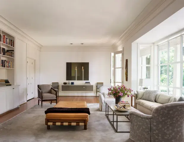 A bright, refined living room with built-in white shelving and cabinetry, a wall-mounted media console, large multi-pane windows, classic crown moulding, and layered seating arranged around a wood coffee table and upholstered ottoman. The room combines traditional architectural detailing with contemporary furniture and a neutral, warm palette.