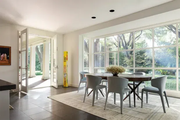 Bright contemporary dining area with a large rectangular timber dining table and upholstered grey dining chairs positioned beside expansive white-framed windows. The room has a light neutral palette, pale tiled flooring, recessed ceiling lights, and strong views of leafy greenery outside. Open glazed doors connect the dining area to an outdoor space or entry, while a colourful narrow artwork adds a small accent.