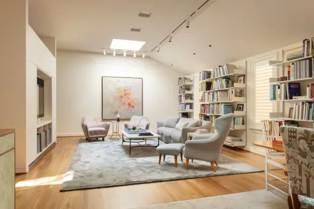 Bright, spacious living room with warm timber flooring, a large neutral area rug, multiple upholstered lounge chairs and sofas arranged around a low coffee table, extensive built-in and freestanding bookshelves, framed artwork, and a television recessed into white joinery. The room has a clean contemporary fit-out, track lighting, a skylight or roof window, and plantation shutters.