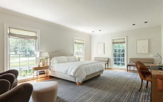 Bright, spacious bedroom with a central upholstered bed, white bedding, light grey walls, broad timber flooring, and several tall windows with woven Roman blinds. The room includes a compact sitting area, a console or desk with a wooden chair, restrained artwork, and traditional ceiling and wall trim. The overall look is calm, refined, and contemporary-classic.
