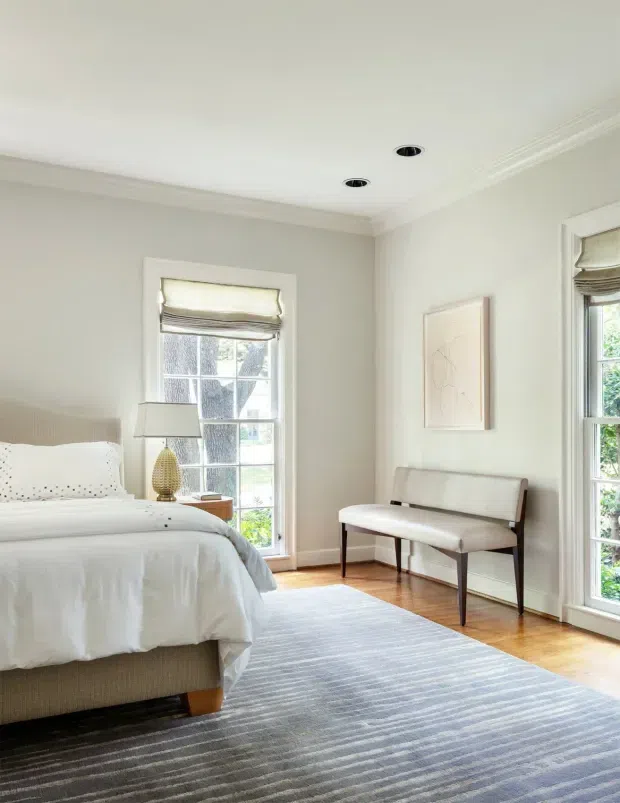 Bright, refined bedroom with a partially visible upholstered bed dressed in white bedding, a pale striped area rug, timber flooring, tall divided-light windows with Roman blinds, and a slim upholstered bench. The room has soft neutral walls, white decorative ceiling moulding, recessed downlights, and abundant natural light.