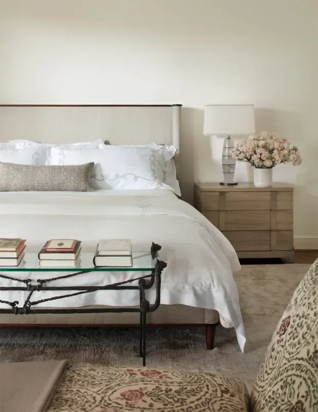A softly styled bedroom featuring a large upholstered headboard with dark trim, layered white bedding and decorative pillows, a timber bedside chest with a lamp and flowers, and a metal-framed bench at the foot of the bed. Neutral carpeting, pale walls, and patterned upholstered seating create a refined, warm interior.