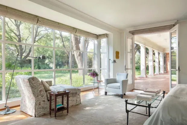 Bright, elegant sunroom or living space with expansive multi-pane windows overlooking a leafy garden. The room features a pale upholstered armchair, a small timber side table, a glass-and-metal coffee table, a floor lamp, crown moulding, and wide open glazed doors leading to a columned verandah or covered terrace. The restrained neutral palette and abundant natural light create a refined, airy atmosphere.