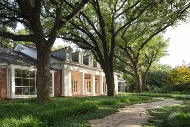 Exterior view of a substantial traditional brick-and-painted residence set beneath mature spreading trees. The facade features a columned portico, tall multi-pane French doors, large divided-light windows, and several dormer windows in a pitched roof. A paved stone walkway curves through a landscaped garden toward the house.